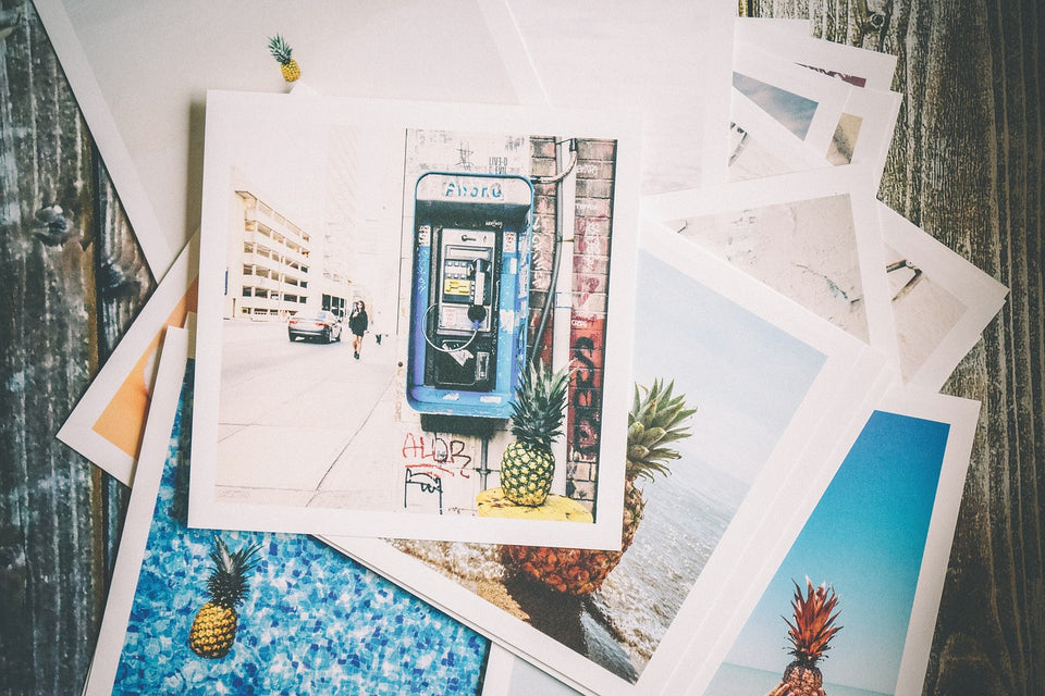 A messy, haphazard stack of photographs of various locales, including a phone booth, a pool with a floating pineapple, and beaches (also with pineapples).